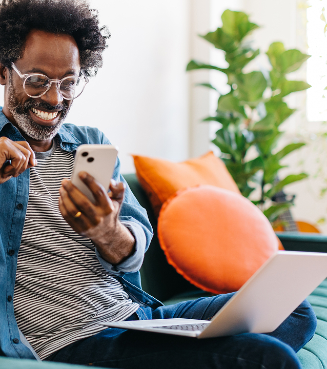 Man sitting on sofa, looking at his phone smiling with a laptop in his lap. Orange pillows and green plant in the background.